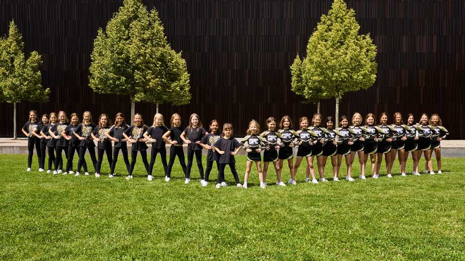 Gruppenfoto von Cheerleadern in einheitlicher Kleidung, auf einer Wiese, vor einem modernen Gebäude mit Bäumen.
