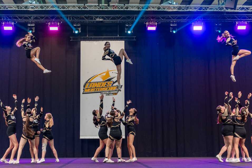 Cheerleaderinnen f&uuml;hren eine akrobatische Pyramide auf, w&auml;hrend eine Sportlerin hoch in die Luft springt.