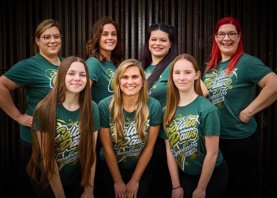 Eine Gruppe von sieben Frauen in grünen T-Shirts posiert lächelnd vor einem dunkel gestrichenen Hintergrund.