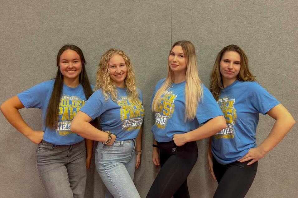 Vier junge Frauen posieren in blauen T-Shirts mit dem Aufdruck „Golden Paws“ vor einer grauen Wand.