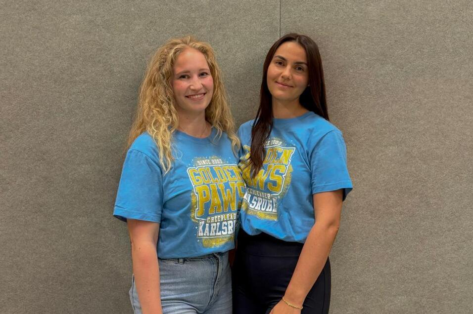 Zwei junge Frauen in blauen T-Shirts stehen lächelnd vor einer grauen Wand, die eine freundliche Atmosphäre schafft.