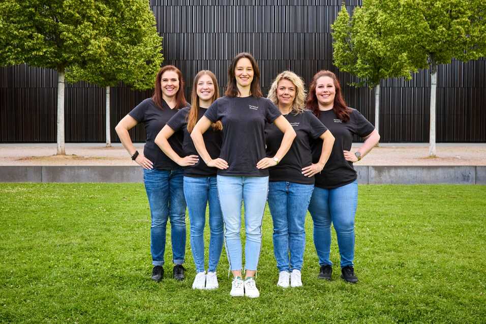 F&uuml;nf Frauen in schwarzen T-Shirts stehen in einer Reihe auf einer Wiese vor einer moderneren Architektur.