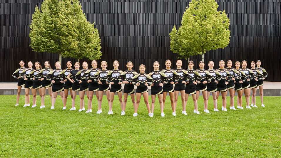 Gruppenportr&auml;t von Cheerleadern in einheitlichen Outfits auf einer gr&uuml;nen Wiese mit B&auml;umen im Hintergrund.