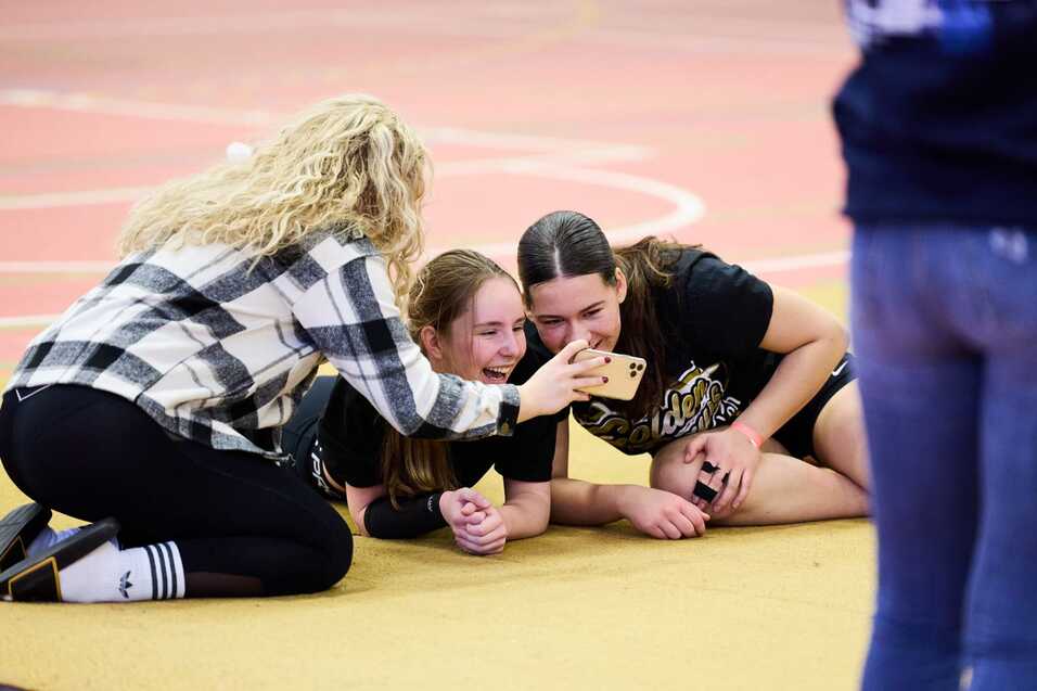 Drei M&auml;dchen sitzen auf einem Sportboden und schauen gemeinsam auf ein Smartphone, w&auml;hrend sie lachen.