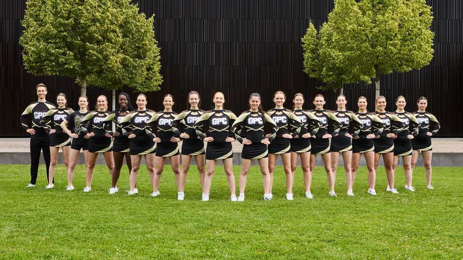 Gruppierung von Cheerleadern in uniformierten Outfits, auf einer Wiese vor einer modernen, strukturierten Wand.