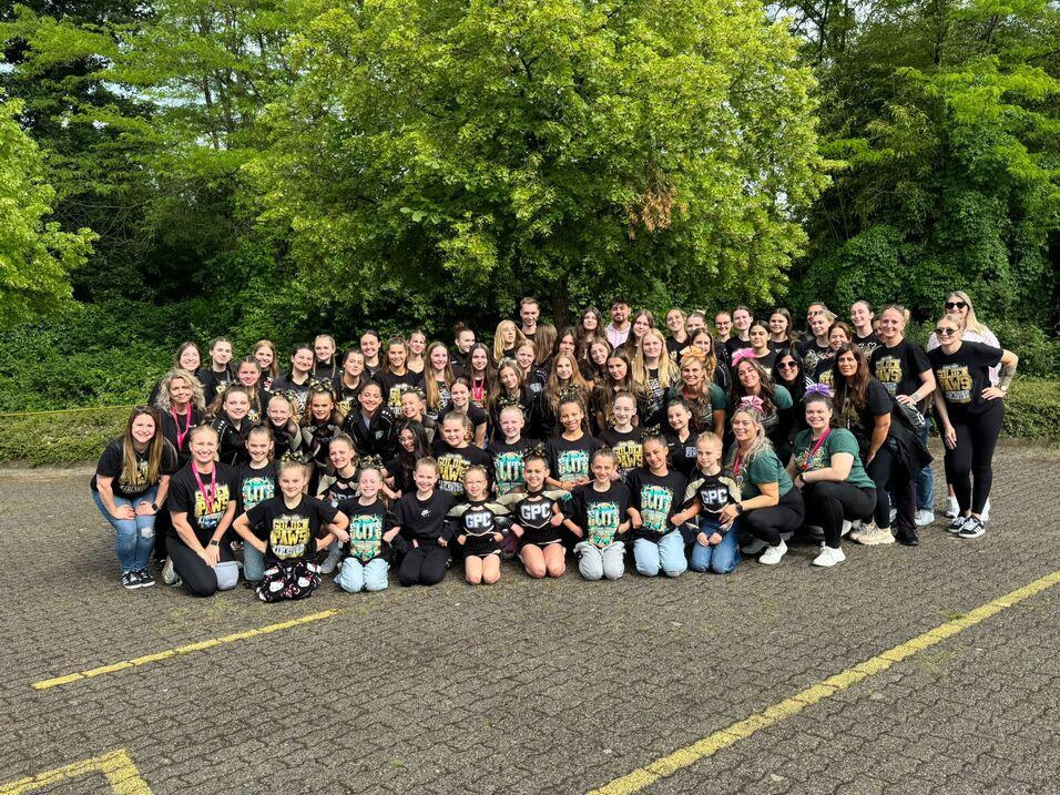 Gruppenszene mit &uuml;ber 70 Cheerleadern und Trainern in einheitlichen T-Shirts vor gr&uuml;ner Landschaft.