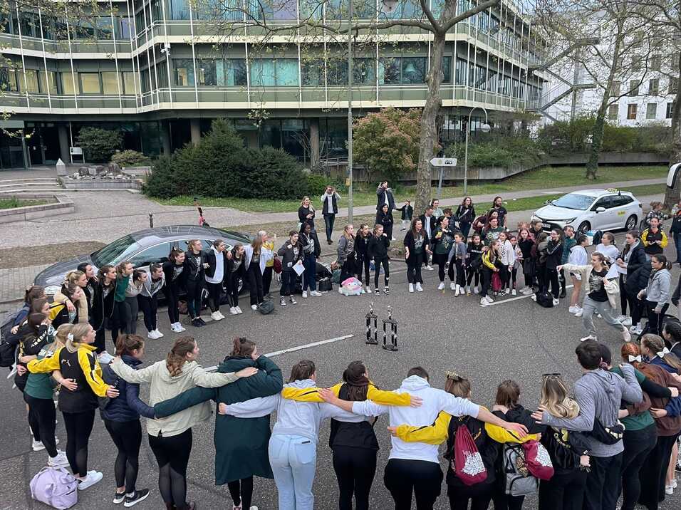 Gruppe von T&auml;nzern in einer Umarmung auf einer Stra&szlig;e, umgeben von Zuschauern und Troph&auml;en im Hintergrund.